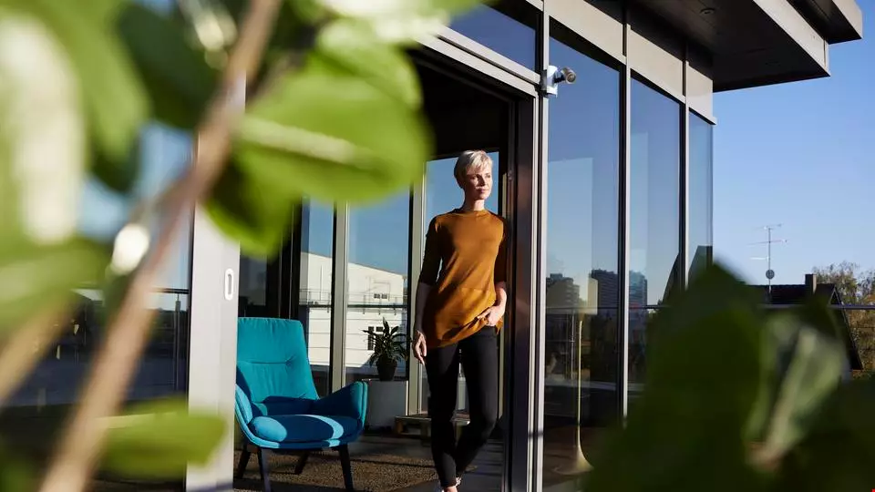 Kvinna i kort hår står avslappnat med ena handen i fickan och tittar ut från en balkongdörr. Det är soligt väder och blå himmel. Det står en blå fåtölj bakom henne inne i lägenheten och i förgrunden är det grön växtlighet.