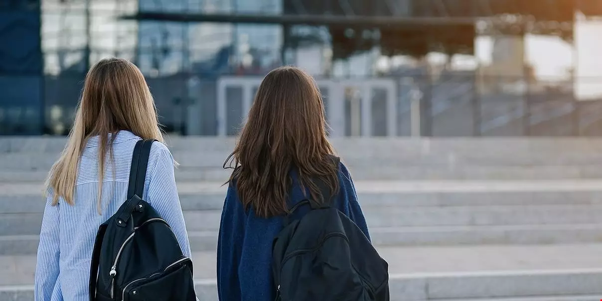 Två unga studenter går tillsammans mot en byggnad med en trappa framför. De har båda svarta ryggsäckar. 