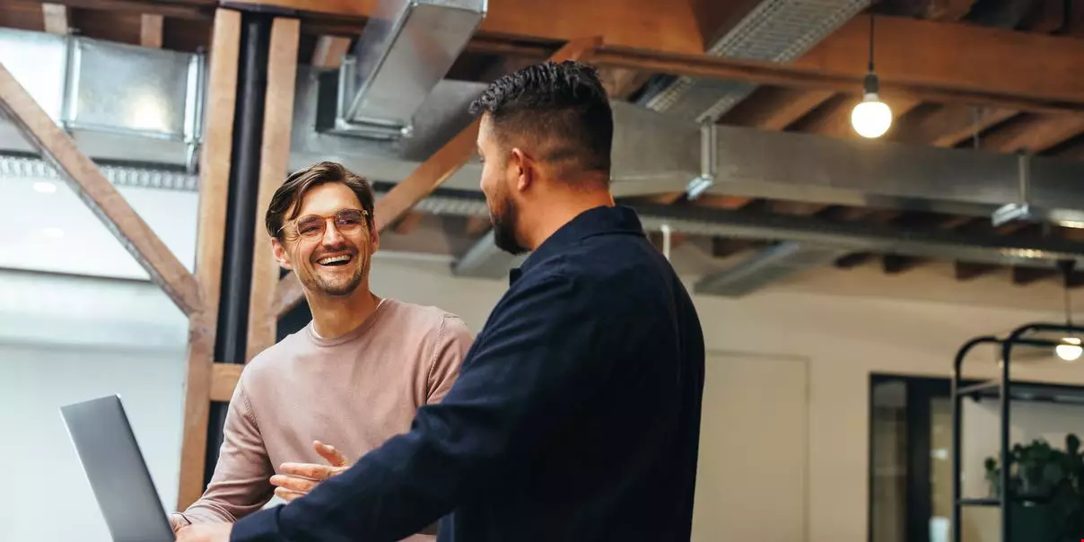 Två manliga kollegor står tillsammans på en arbetsplats och skrattar ihop.